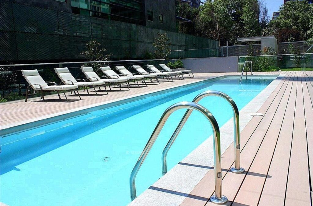 Piscina del edificio con reposeras, deck de madera y áreas verdes en condominio de Callao 3860, Las Condes.